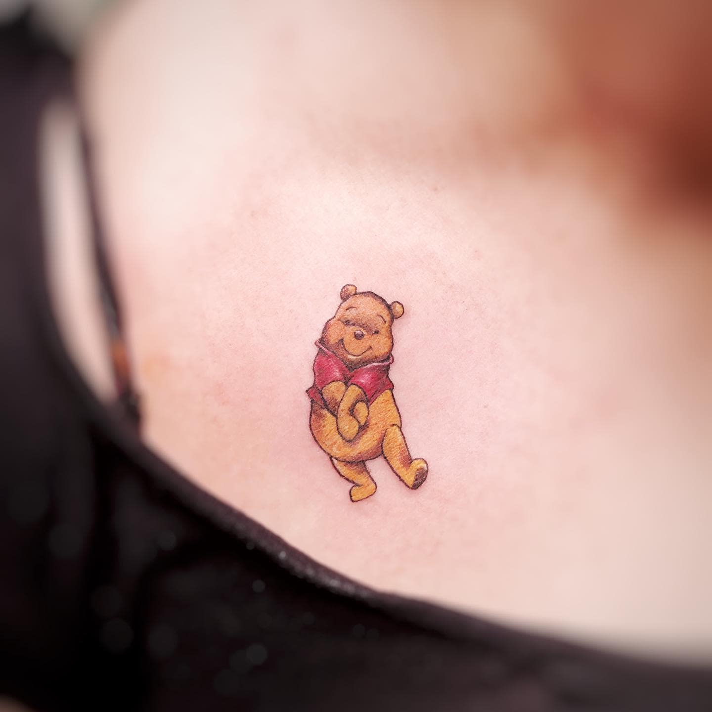 Small color tattoo of Winnie the Pooh standing with a happy expression on a person's shoulder